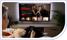 Person holding popcorn and a remote, watching a TV screen showing a comedy series with three women in elegant outfits, set in a cozy living room.