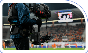 Camera operator with professional gear films a soccer game. Blurred stadium and energetic crowd in the background convey excitement.